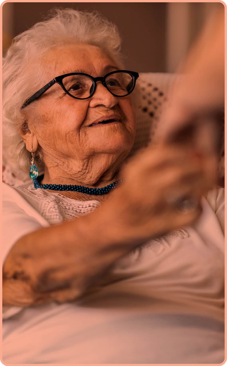 Female home carer supporting old woman to stand up from the armchair at care home