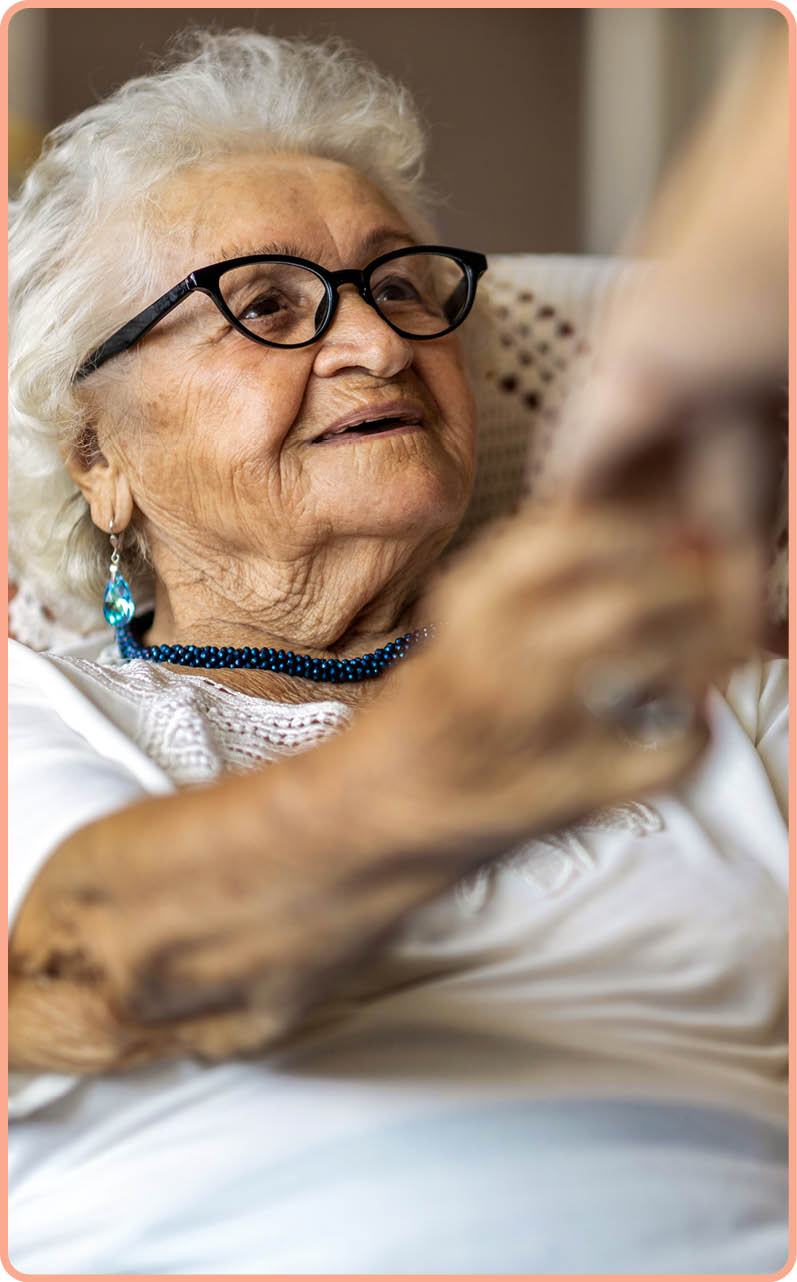 Female home carer supporting old woman to stand up from the armchair at care home